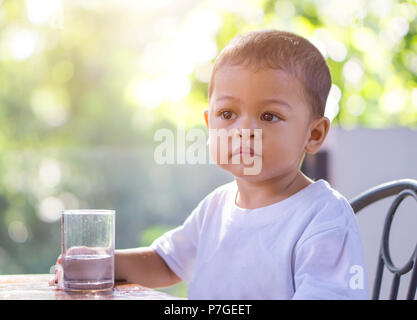 Little asian baby going to drink water in a glass cup in the morning time with nature bokeh background. Stock Photo