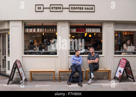 Two male friends sitting and talking on wooden bench outside of 39 Steps Coffee Haus, London, UK Stock Photo