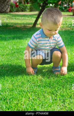 Cute Caucasian Blond Baby Boy with Blue Eyes Toddler of 2 Years Old Sits on Haunches on Green Grass in City Park Playing. Vivid Bright Summer Day Gree Stock Photo