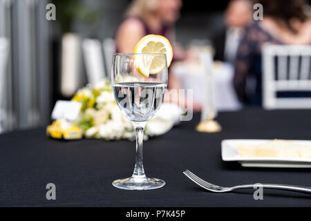 Closeup of one glass with lemon slice and water in restaurant or wedding reception black table tablecloth, bokeh of people, plate with cake slice, bou Stock Photo