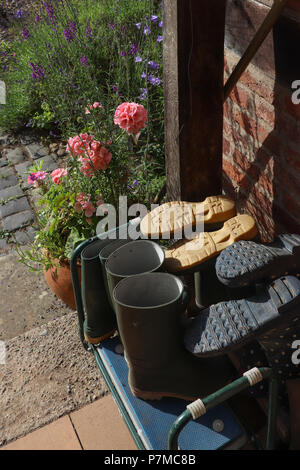 Several pairs of a gardener's wellie boots stacked on an entry porch by a red brick wall; pink geraniums, lavender and blue bells in garden nearby. Stock Photo