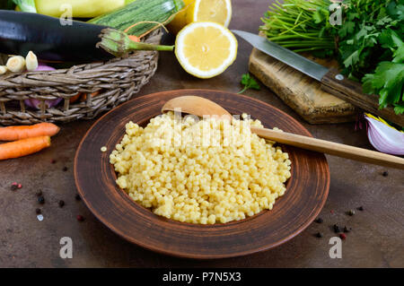 Delicious useful porridge cous cous in a clay plate and raw fresh vegetables in a basket, greens, lemon. Ingredients for cooking Tabbouleh. Vegetarian Stock Photo