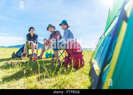 Group of travelers camping and doing picnic in meadow with tent foreground. Mountain and lake background. People and lifestyles concept. Outdoors acti Stock Photo