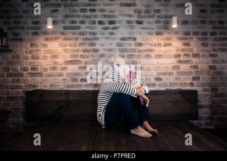 Unusual young man in comical mask sits alone on bed in stylish apartment. Sad unicorn in casual clothes. Freaky man after break up spend time at home Stock Photo