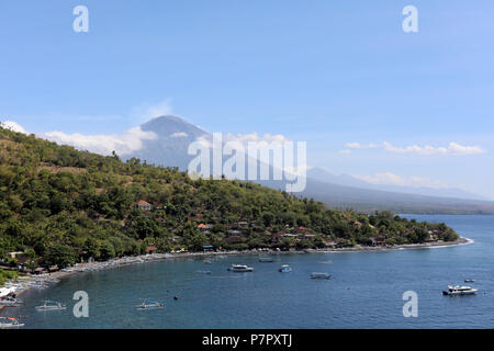 Amed, Indonesia – July 1 2018: View of the smoking volcano of Mount Agung in east Bali, Indonesia, with the black sand beach of Jemeluk in the foregro Stock Photo