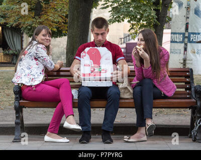 LVIV, UKRAINE - AUGUST 19, 2015: Three youngsters, teenagers, one caucasian male and two females, eating pizza and smiling sitting on a bench in downt Stock Photo