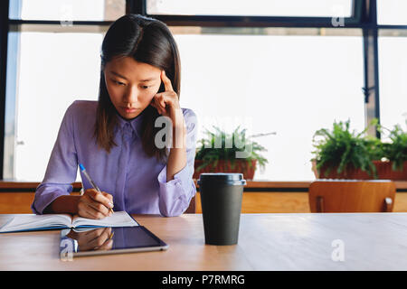 Asian female student sitting in cafe and writing on notebook, looking at digital tablet Stock Photo