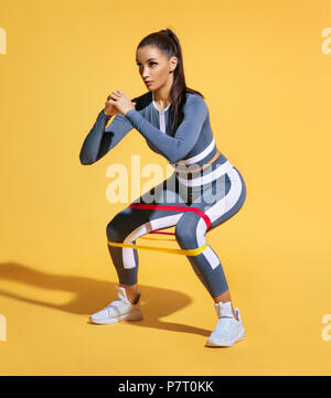 Sporty woman squatting doing sit-ups with resistance band. Photo of latin woman in fashionable sportswear on yellow background. Strength and motivatio Stock Photo