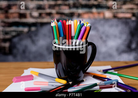 Art Markers and Colored Pencils in a Coffee cup.  Below the cup on a wooden desk is paper and scattered Markers and Pencils  All against a vintage Bri Stock Photo