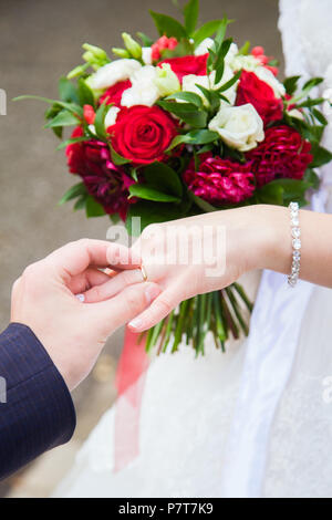 bride puts on a gold ring on the bridegroom's hand, on a background of a white dress and a floral bouquet Stock Photo