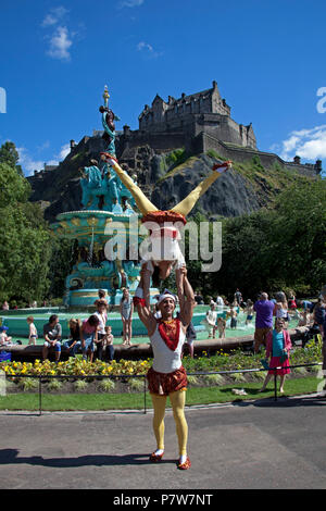 Edinburgh, Scotland, UK. 8 July 2018. Ross Fountain, West Princes Street Gardens switch on day, crowds of families and tourists flock in the sunshine to see the circus acts and Edinburgh School's pipe band and bathe in the fountain water which was switched off in 2010. Stock Photo