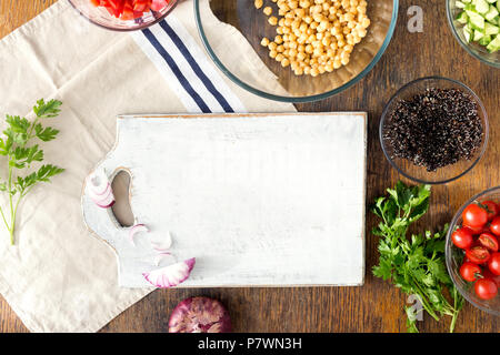 Around the wooden kitchen board, black quinoa, chickpeas, peppers, onion, cucumber, tomatoes and parsley on rustic wooden table, top view. Ingredients Stock Photo