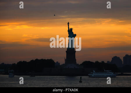 The Statue of Liberty silhouetted against a glowing bright orange/yellow sky on a spring evening, as seen from the Staten Island Ferry; 3 boats, bird. Stock Photo
