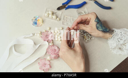 Girl fashion designer makes jewelry. Shooting close-up. She pushes the artificial pearls on the pin. Nippers, round-nosed pliers lie on the table Stock Photo