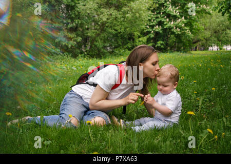 Family. Son and mother outdoors. A pregnant girl with a backpack is sitting on the lawn and kissing a little boy. He sits on the lawn and plays with a Stock Photo