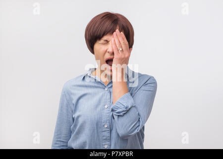 Portrait of mature woman in blue shirt, yawning with her eyes closed and covering her mouth, isolated over white wall. Stock Photo