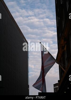 Silhouette of Union Jack (British) flag flying between the buildings against dark blue sky with clouds. Stock Photo