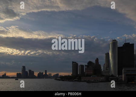 Dramatic sky at dusk over the New Jersey/New York harbor where the Hudson River begins. Large blue opening in gray sky above silhouetted skylines. Stock Photo