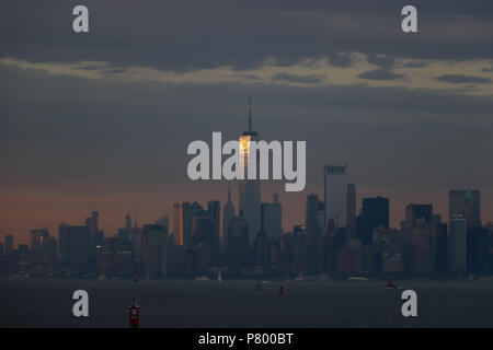 The last rays of a setting sun catch the top of One World Trade Center on the New York City skyline, just visible across the harbor at evening dusk. Stock Photo