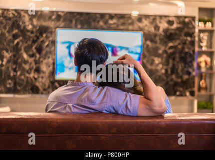 Asian young lovers watching television on sofa. Couples and Relax concept. Holiday and vacation concept. Night dating and honeymoon theme. Back view Stock Photo