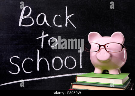 Pink piggy bank with glasses standing on textbooks in front of a blackboard with back to school message. Stock Photo