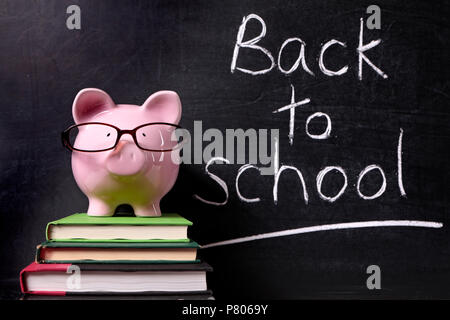 Pink piggy bank with glasses standing on textbooks in front of a blackboard with back to school message. Stock Photo