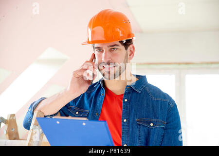 Portrait of young handyman holding clipboard in his hand and making call while standing at construction area. Stock Photo