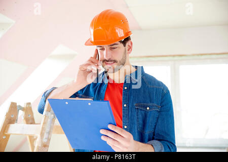 Portrait of young handyman holding clipboard in his hand and making call while standing at construction area. Stock Photo