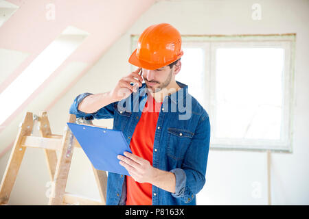 Portrait of young handyman holding clipboard in his hand and making call while standing at construction area. Stock Photo
