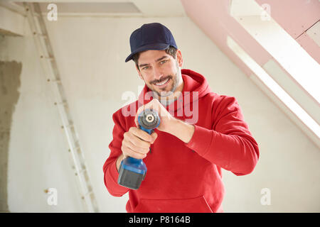 Portrait of a smiling young repairman wearing baseball cap and tool belt while standing at construction site and holding power drill in his hand. Stock Photo