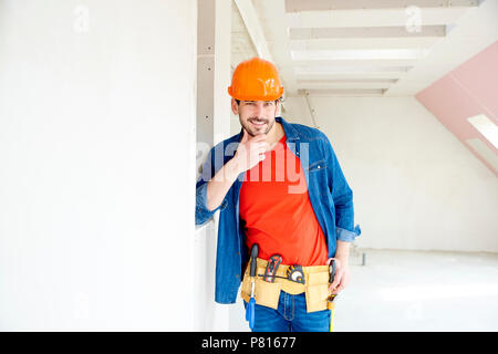 Portrait of handsome young handyman wearing hard hat and tool belt while standing in the house. Stock Photo