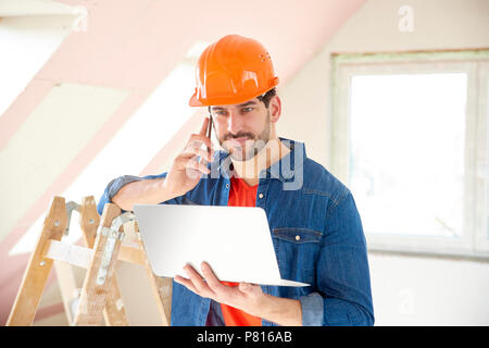 Young repairman using laptop and mobile phone while standing at renovation area. Stock Photo