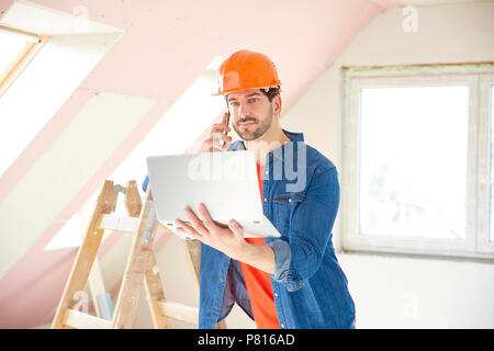 Young repairman using laptop and mobile phone while standing at renovation area. Stock Photo