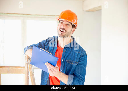 Portrait of young handyman wearing hard hat and holding clipboard in his hand while standing at construction area. Stock Photo
