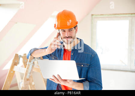 Young repairman using laptop and mobile phone while standing at renovation area. Stock Photo