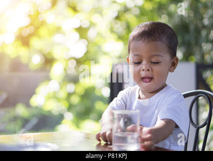 Little asian baby going to drink water in a glass cup in the morning time with nature bokeh background. Stock Photo