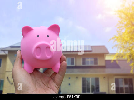 Male hand holding a pink piggy bank on house, real estate or home blured background. Stock Photo
