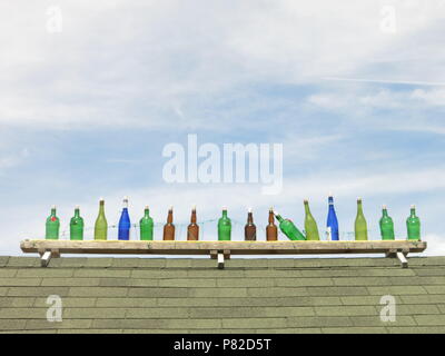 Ten green bottles (plus a few others) standing in a row on a wall against a blue and cloudy sky; and the nursery rhyme says one began to fall Stock Photo