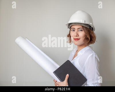 Beautiful asian woman engineer curly short hair wearing white safety helmet and white shirt smiling and holding blueprint and notebook on white wall b Stock Photo