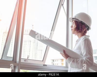 Beautiful asian woman engineer curly short hair wearing white safety helmet and white shirt holding blueprint near the glass window with construction  Stock Photo