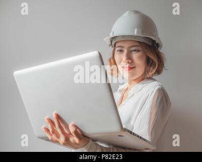 Beautiful asian woman engineer curly short hair wearing white safety helmet and white shirt working with silver laptop and smiling. Working woman conc Stock Photo