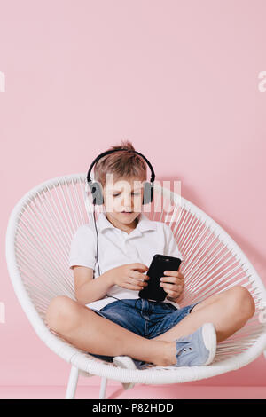 Adorable child in headphones sitting with crossed legs on white woven round chair and concentrating on mobile phone in hands on pink background Stock Photo