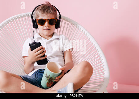 Cute happy relaxed kid in summer clothes and headphones sitting with crossed legs on round woven chair and using mobile phone over pink studio backgro Stock Photo