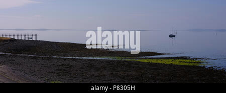 Panoramic view across Strangford Lough in summer mist and clear reflections of clouds and boats on the horizon. A wooden jetty frames the picture. Stock Photo