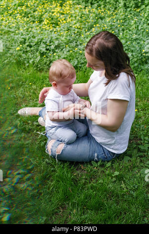 Family. Son and mother outdoors. Pregnant girl sits on lawn and holds small boy in her lap. He plays with her bracelet. Blur effect. Stock Photo