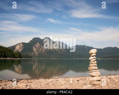 Balanced stone pyramide on shore of blue water of mountain lake. Blue mountains in water level mirror. Stock Photo