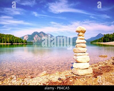 Balanced stone pyramide on shore of blue water of mountain lake. Blue mountains in water level mirror. Stock Photo