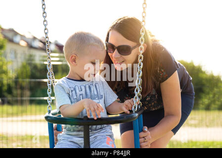 Happy mother with baby sits on the swing. Leisure time on the playground Stock Photo