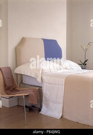 Bed with a cream+blue upholstered head board and white pillows and sheet in an economy style bedroom with a metal+wicker chair Stock Photo