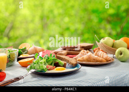 Breakfast table with fried eggs, croissants, fruit, salad and juice on a background of green leaves bokeh Stock Photo